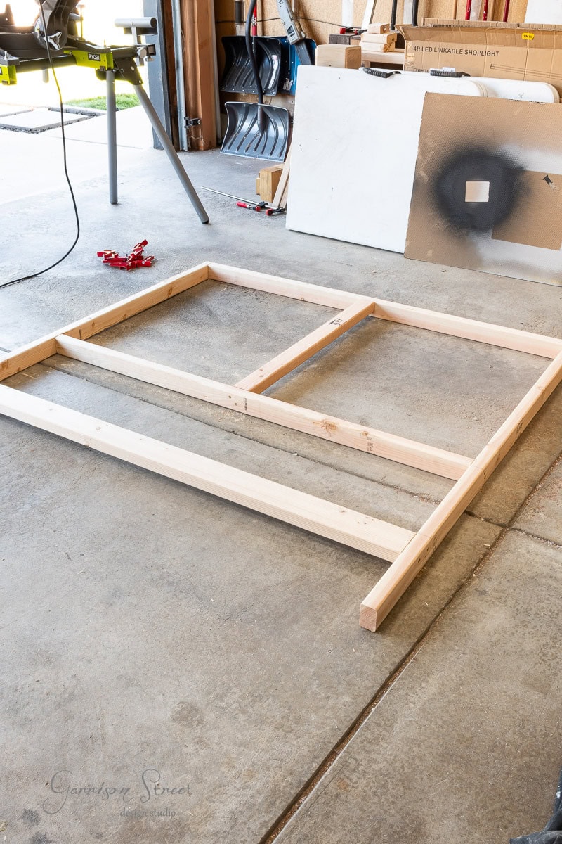 A wooden frame, positioned to eventually become a queen headboard, is laid on a concrete floor in a garage workshop. Tools and materials are visible in the background, including a table saw, spray-painted boards, and clamps—ready for this DIY project.