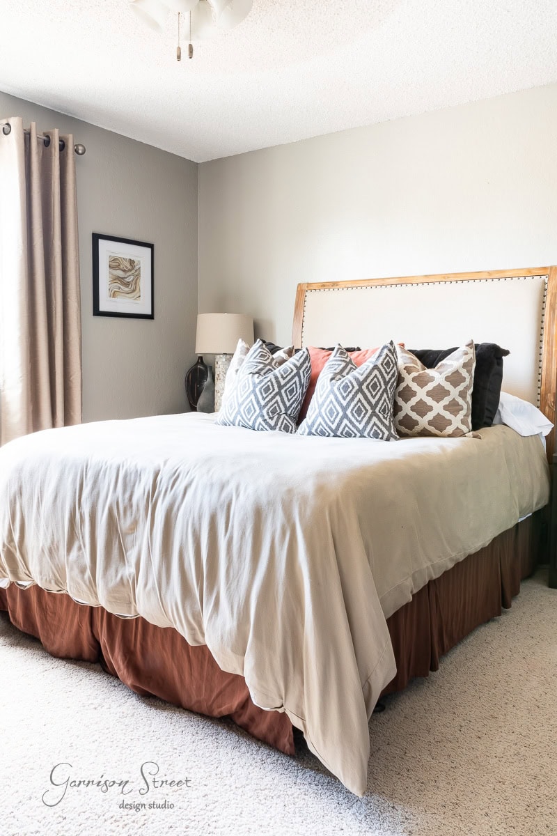 A cozy bedroom showcases a large queen bed with patterned cushions against a padded DIY headboard. Neutral tones are used throughout, with curtains hanging by the window and framed artwork adorning the wall. A lamp sits on the bedside table, casting a warm glow.