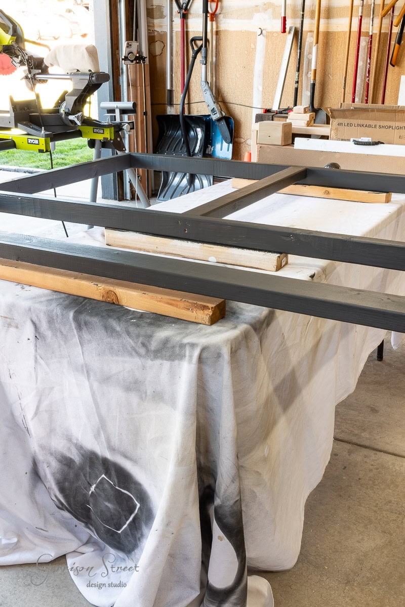 In a bustling workshop, a DIY headboard is being painted black. The queen-size wooden frame is propped up on blocks over a table draped with a drop cloth. Surrounding this scene are various tools, including a miter saw, all contributing to the creative frenzy.