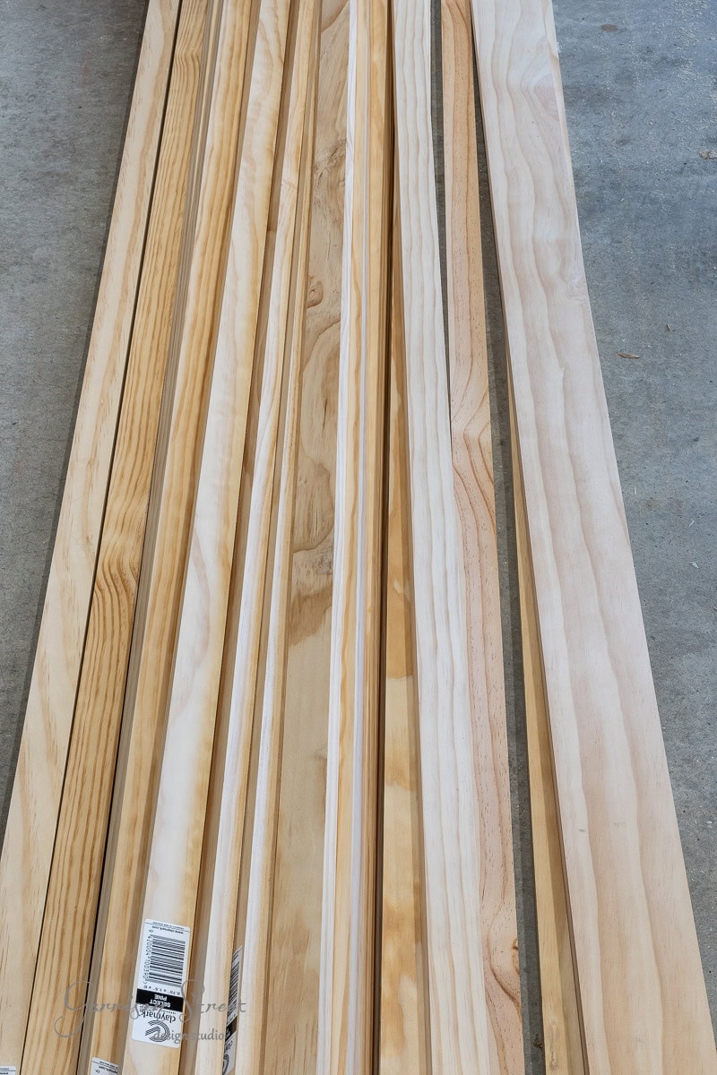 Long planks of wood are neatly arranged on a concrete surface, perfect for a DIY headboard project. The wood varies in shades of brown, with visible grain patterns, ideal for creating a unique queen-sized piece. Some planks have stickers on one end.