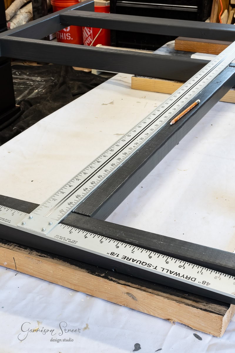 A close-up of a wooden tabletop with a black painted frame hints at a DIY project, possibly crafting a headboard. A large metal T-square ruler and pencil rest on the frame, suggesting precise measurement work. Various tools and materials are scattered in the background, ready for use.