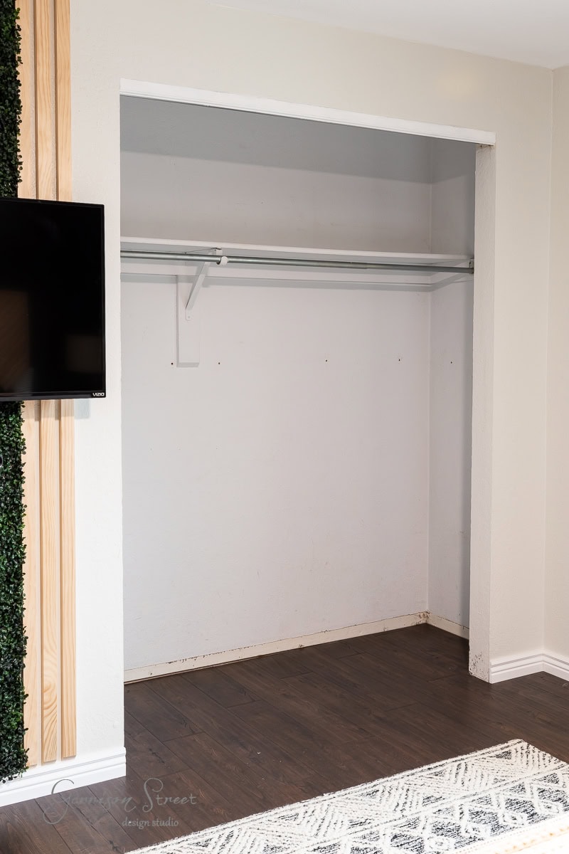 A vacant closet with a white interior and a single metal rod for hanging clothes awaits a DIY makeover. The floor is dark wood, and a section of a wall-mounted TV is visible on the left, complemented by a narrow strip of vertical wood paneling.