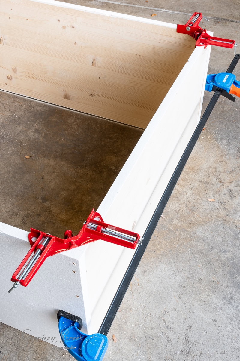 A wooden box frame is being assembled on a concrete floor for a DIY closet makeover. Red and blue clamps secure the corners and edges, holding the pieces together tightly. Small bits of sawdust speckle the area around the box.