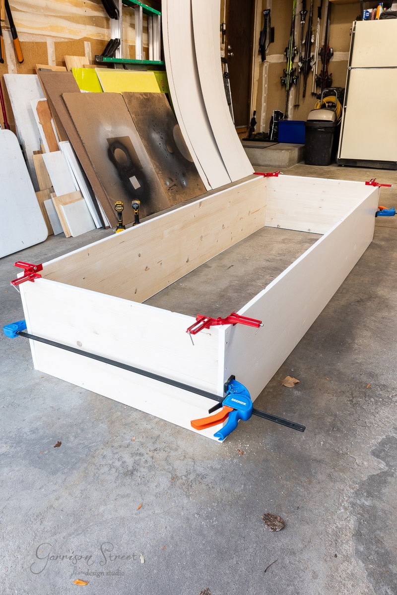 A partially assembled wooden rectangular frame, destined for a closet makeover, is secured with red and blue clamps in a garage. Various tools and materials, essential for this DIY project, are visible in the background, including panels and a broom.