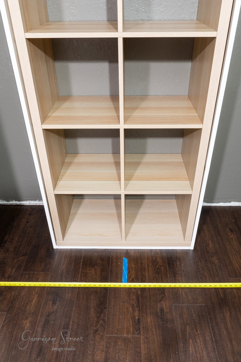 A wooden bookshelf with eight compartments stands on a dark hardwood floor, part of a closet makeover. A yellow measuring tape is laid out in front, showcasing measurements for this DIY project. A small blue tape mark is visible on the floor at the base of the bookshelf.
