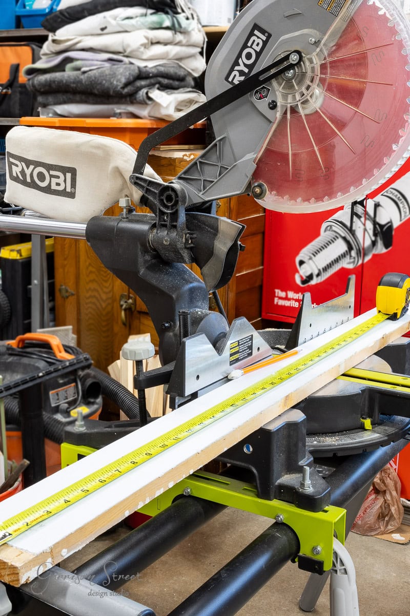 A Ryobi mitre saw is set up on a workbench, ready to bring your closet makeover DIY to life. A piece of wood rests under the blade, measured precisely with a tape laid along it. Various tools and equipment are scattered in the background, each poised to play its part in your project.
