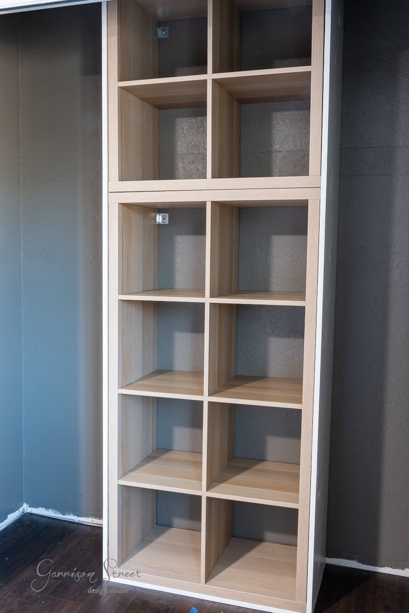 A tall, light wooden bookshelf with multiple square compartments rests against a dark wall, reminiscent of a DIY closet makeover. The bookshelf's simple, modern design and its empty shelves invite creativity and personalization.