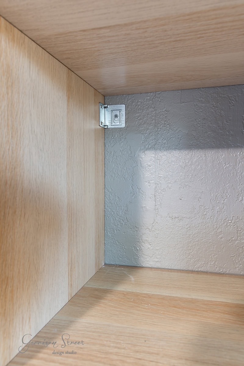 The image shows the interior corner of a wooden cabinet, reminiscent of a DIY closet makeover. A metal bracket is attached to the top corner, securing the panel. The walls boast a textured gray finish, while the wood has a light finish.