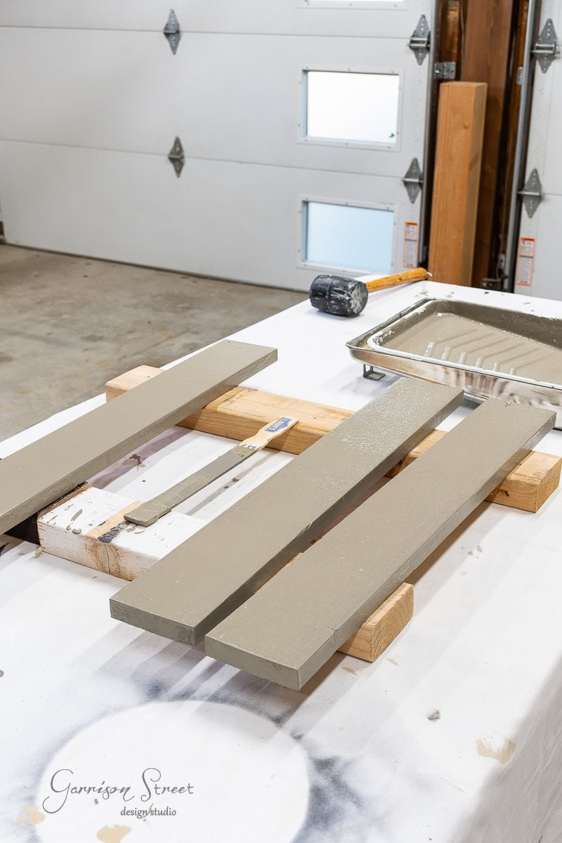 Two wooden boards on a worktable are painted taupe, lying parallel on wooden supports. As part of a closet makeover DIY project, a paint tray with taupe paint and a roller sits to the right, next to a rubber mallet. The background features a garage door.