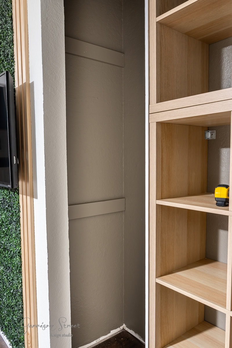 A small, empty closet with a wooden frame and open shelving unit on the right awaits your DIY touch. The wall on the left is adorned with green foliage, offering inspiration for a closet makeover. A yellow and black tape measure rests on the shelf, ready to help you transform the space.