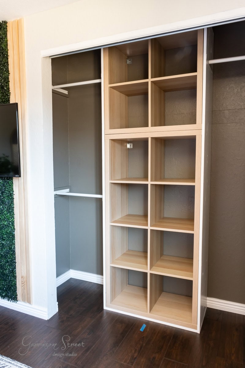A wooden closet organizer with multiple open shelves is installed in a partially open closet, showcasing a stunning DIY closet makeover. The floor is wooden, and there is a section of green, plant-like wall decoration on the left.