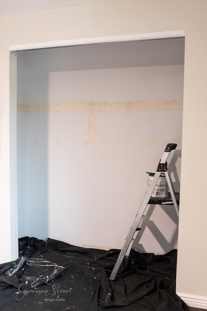 The empty closet, primed for a DIY makeover, features unfinished walls and a step ladder with a paint can poised on top. A black cloth drapes the floor, evoking the excitement of ongoing renovations.