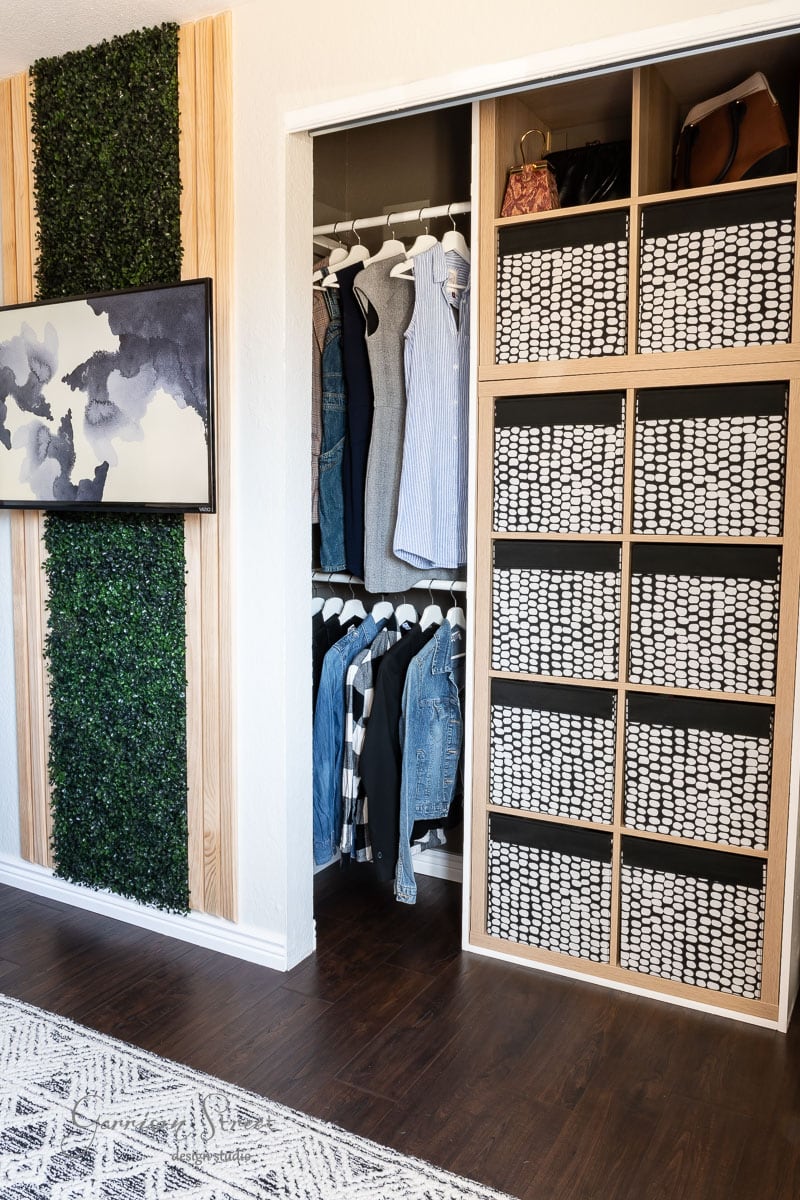 A modern closet makeover showcases a wooden organizer with eight patterned baskets on the right and clothing on black hangers to the left. Vertical greenery and abstract art adorn the wall, while a patterned rug enhances the dark wooden floor, adding a DIY touch of style.