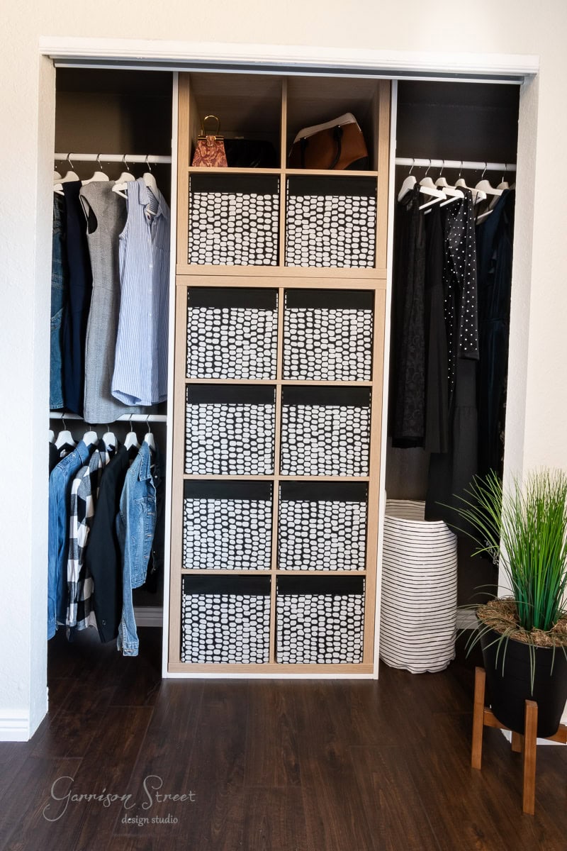 A neatly organized closet makeover features hanging clothes on each side and a central shelving unit filled with matching patterned storage bins. A potted plant and a woven basket are placed on the dark wooden floor, showcasing the DIY transformation's charming touch.