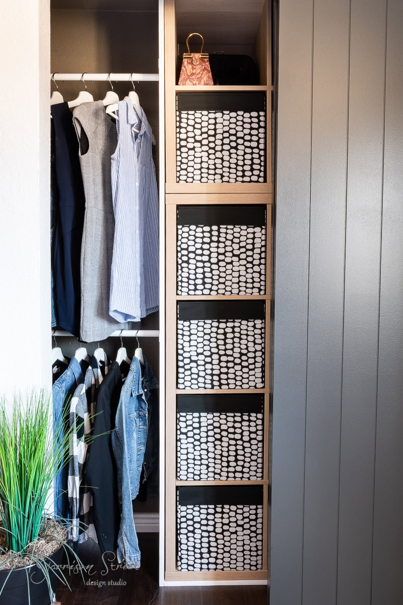 A modern closet makeover showcases neatly arranged clothing on hangers, including dresses and jackets. Adjacent are four shelves with checkered storage bins, a testament to clever DIY organization. A small black handbag rests on the top shelf, while a potted plant adds a touch of greenery in the corner.