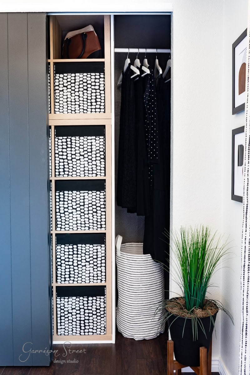 A modern closet makeover with a sliding door showcases shelves of black and white storage baskets. Hanging clothes include black and white items, adding to the chic aesthetic. Below sits a white laundry basket, while a green potted plant adds vibrancy on the right. Perfect for a DIY enthusiast!.