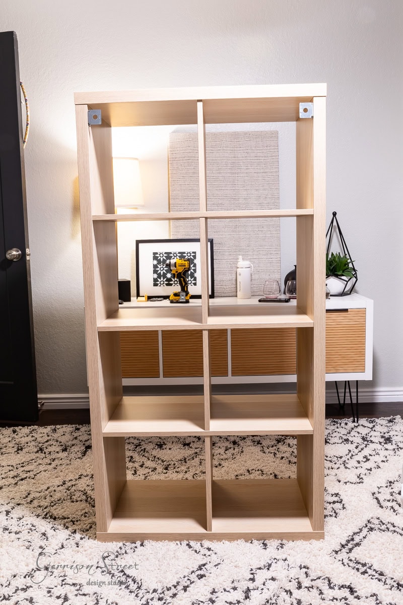 A wooden bookshelf with nine open compartments stands in a room transformed by a closet makeover. Behind it, a white console table with a wooden front holds various decor items. The space, invigorated by DIY touches, features a textured rug, a lamp, and decorative plants.
