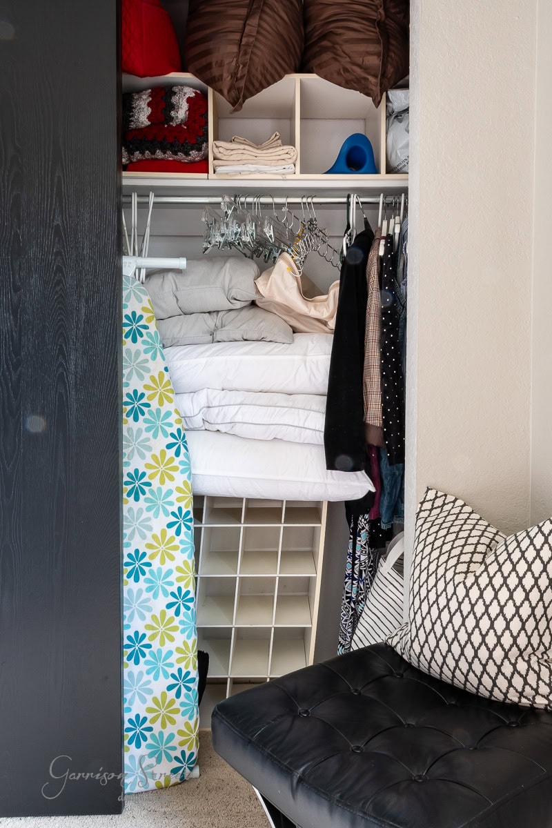 A closet makeover showcases neatly folded blankets, pillows, and hangers organizing clothing. An ironing board with a floral pattern leans against the wall. Completing this DIY transformation, a padded black chair with patterned cushions is positioned in front of the closet.