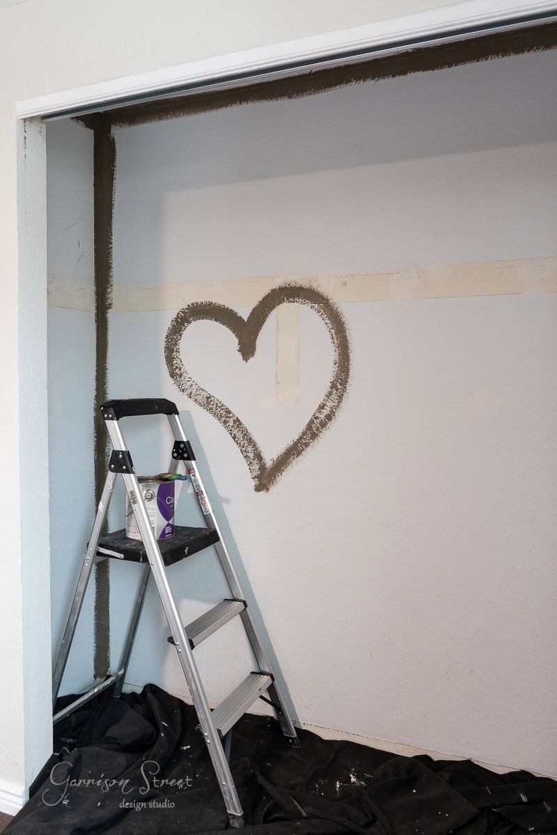 A heart shape is painted on a wall with a dark border, hinting at the start of a closet makeover. A ladder stands in front, with a paintbrush on one of the steps and a black cloth covering the floor below.