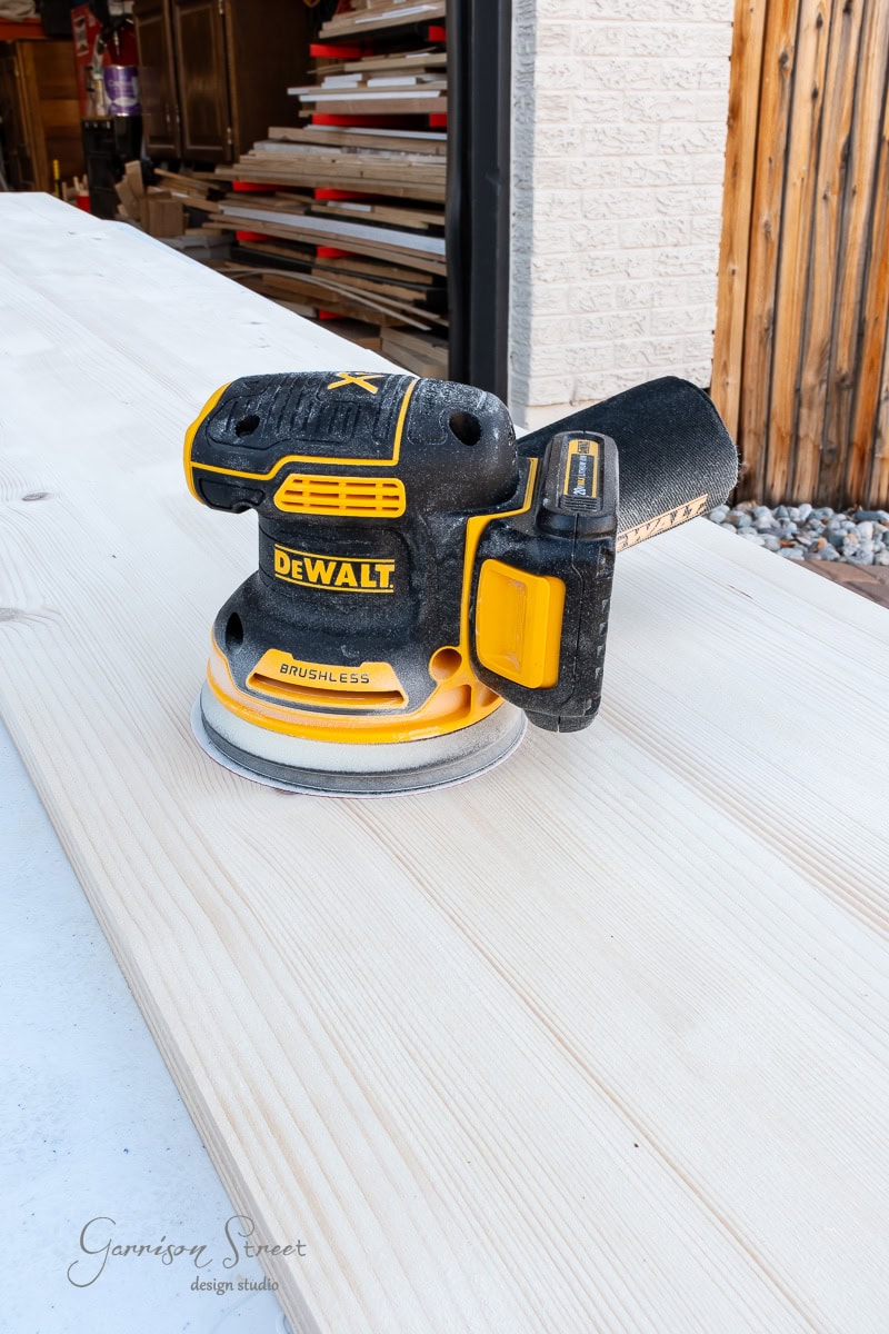 A DeWalt orbital sander rests on a wooden plank, ready for your closet makeover DIY project. The black and yellow tool, with its visible branding, is positioned on a smooth, light-colored surface. Stacks of wood and a stone wall provide the perfect backdrop for your home renovation journey.