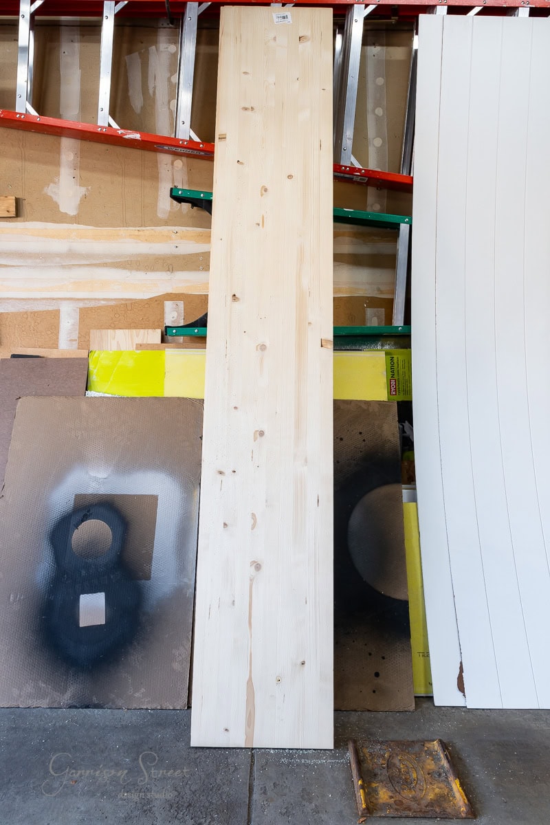 A large wooden board leaning against a garage wall, surrounded by various materials and tools for a closet makeover DIY project. The wall displays a ladder, spray paint art, and other construction items. The floor features painted boards and a yellow caution sign.