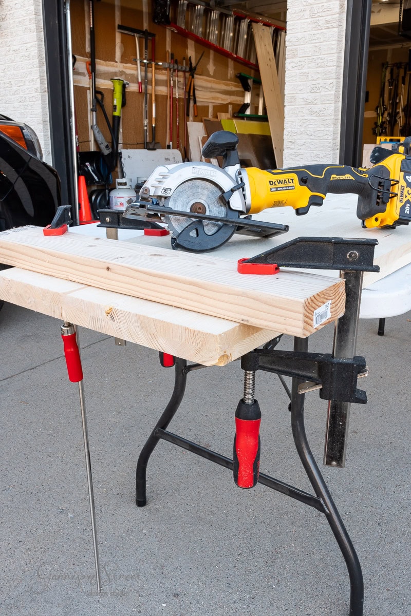 A circular saw rests atop wooden boards clamped to a workbench in a garage, ready for the next DIY project. The garage is filled with various tools and equipment as sunlight illuminates the setup, perfect for planning a closet makeover.