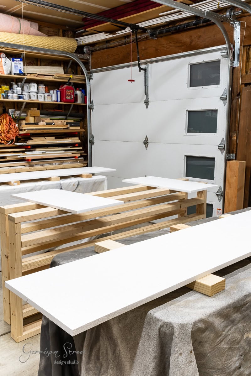 A garage workshop with wooden workbenches holding large white boards, perfect for closet makeover projects. Shelving units brimming with tools and supplies stand ready for any DIY endeavor. A partially open garage door bathes the space in natural light, setting the stage for creativity.