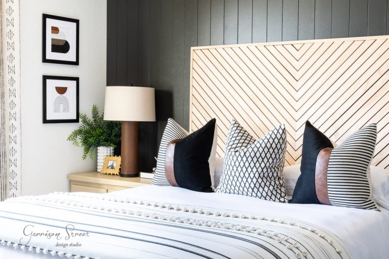 A modern bedroom features a chevron-patterned headboard, four decorative pillows, and a striped blanket. A bedside table with a lamp and plant complements the space, while two framed geometric prints and printable boho art bring character to the dark, textured wall.