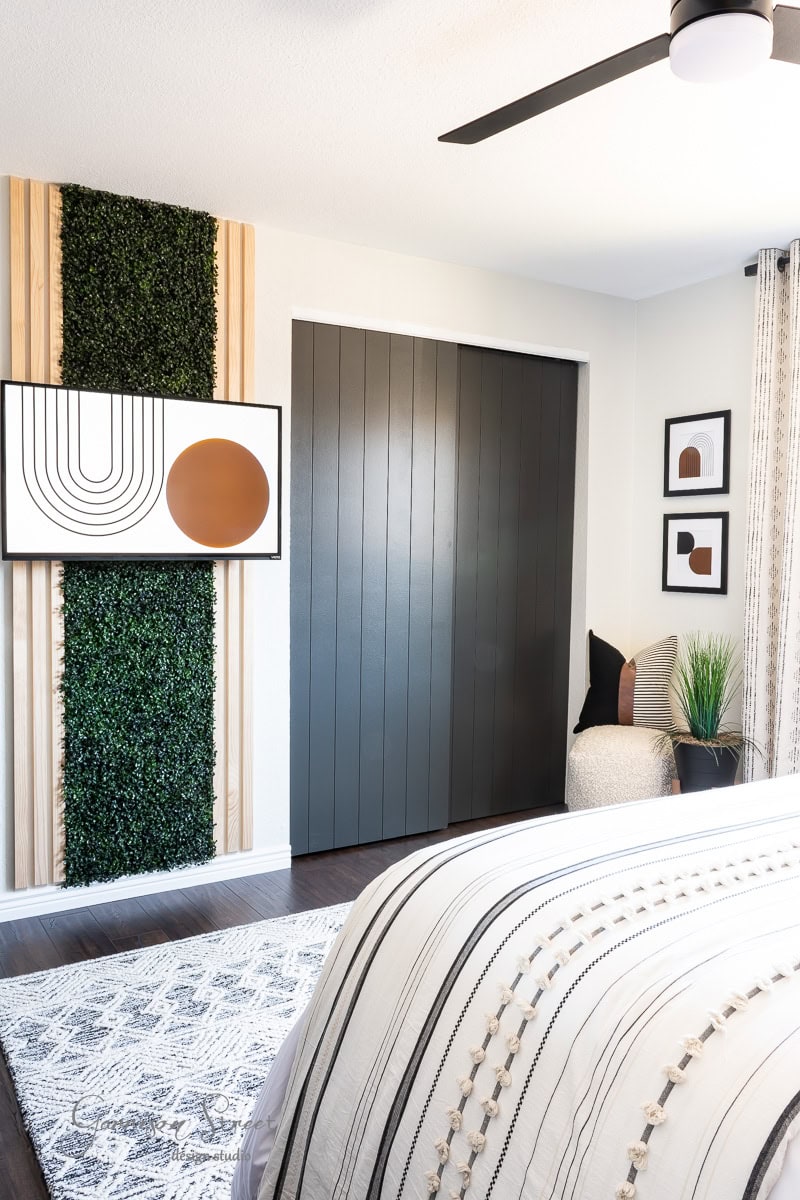 Modern bedroom with a black paneled accent wall and abstract art featuring geometric shapes. The bed has a striped, textured comforter. A gray rug covers the floor, complemented by printable boho art on the walls. A plant beside a cozy chair adds greenery, while a ceiling fan is overhead.