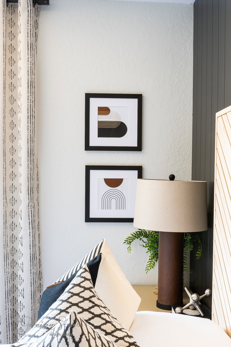 A modern bedroom corner showcasing geometric and printable boho artwork, a black-and-white patterned cushion, and a taupe lamp on a wooden nightstand. The wall behind is partially striped, with a potted plant cozily nestled beside the lamp.
