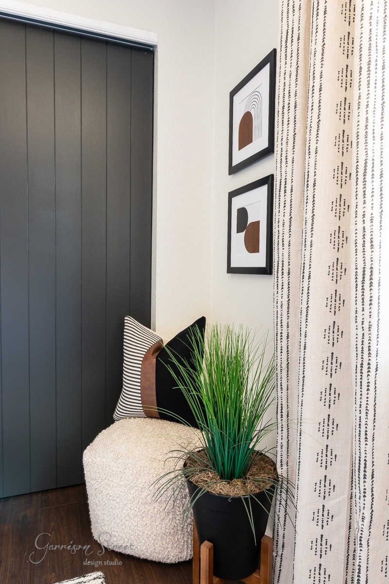 A cozy corner features a fluffy round stool with striped and patterned throw pillows. A tall, green potted plant sits beside it. Two pieces of printable boho art hang on the wall above, and a patterned curtain is on the right.