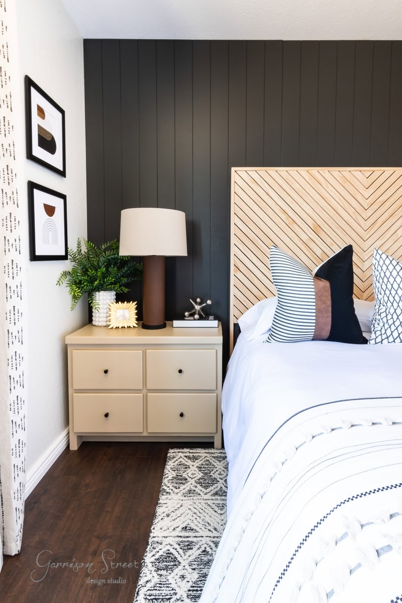A modern bedroom features a bed with white and patterned pillows, a black accent wall, and a wooden headboard. A beige nightstand holds a lamp and plant, while framed printable boho art adorns the walls. A textured rug covers the dark hardwood floor.