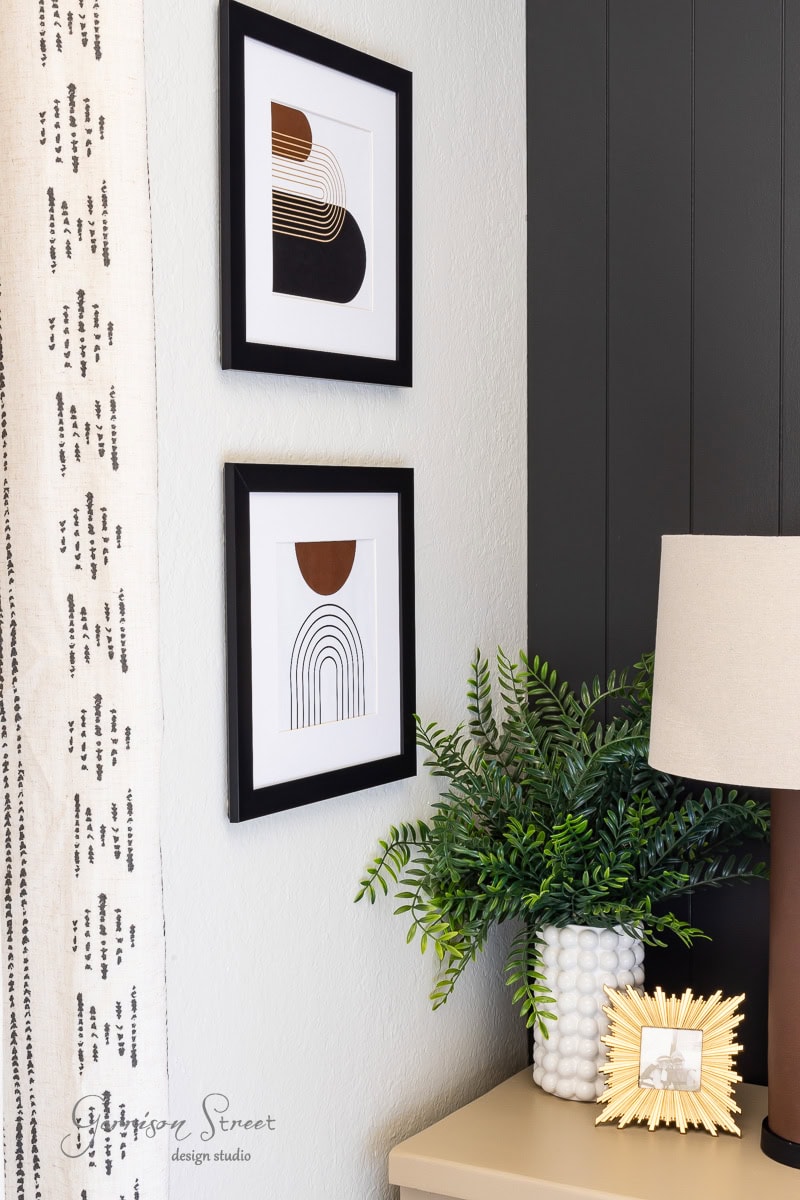 A stylish interior corner features two abstract framed artworks alongside printable boho art, a lush green plant, a decorative picture frame, and a beige table lamp. The wall's partial white and black design nicely complements the modern decor.