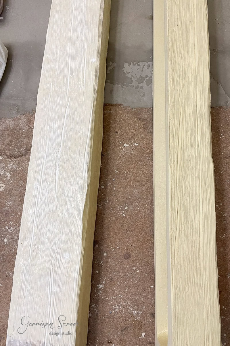 Two elongated pieces of textured wood lie parallel on a surface. One piece is cream-colored, and the other is a lighter beige. The background consists of a mix of rough beige carpet and smooth gray flooring.