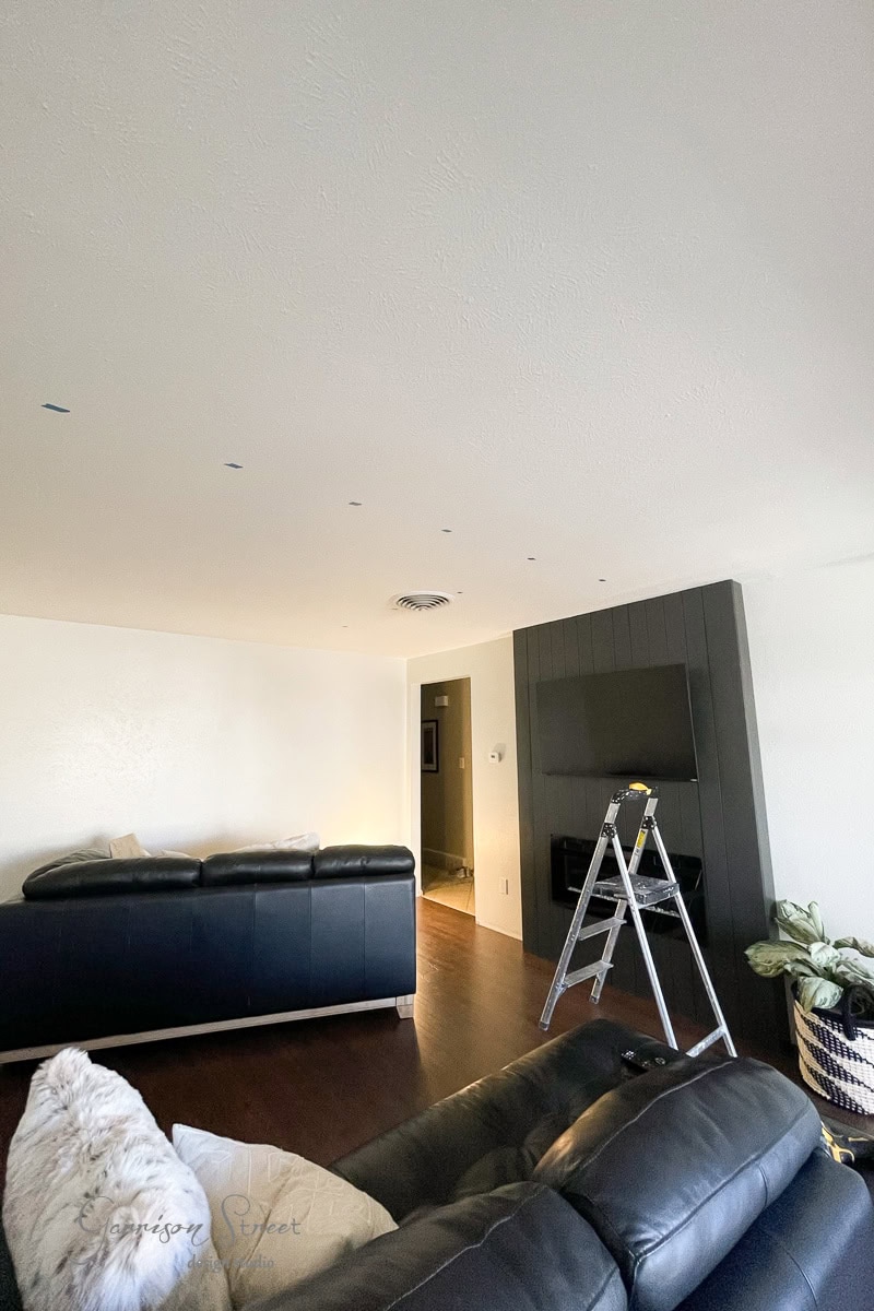 A modern living room features a black couch and a TV mounted above the fireplace. Nearby, a ladder leans against the wall. Dark wood flooring contrasts with the white ceiling, while a plant adorns the corner and a plush blanket rests on the couch.