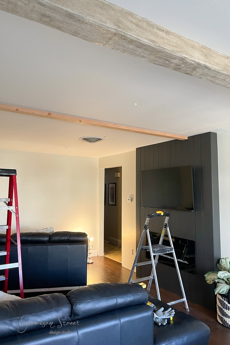 A living room under renovation features a black accent wall with a mounted TV. Ladders and tools indicate ongoing work, with beams partially constructed above. Black leather sofas are positioned in the space, complementing the modern aesthetic.