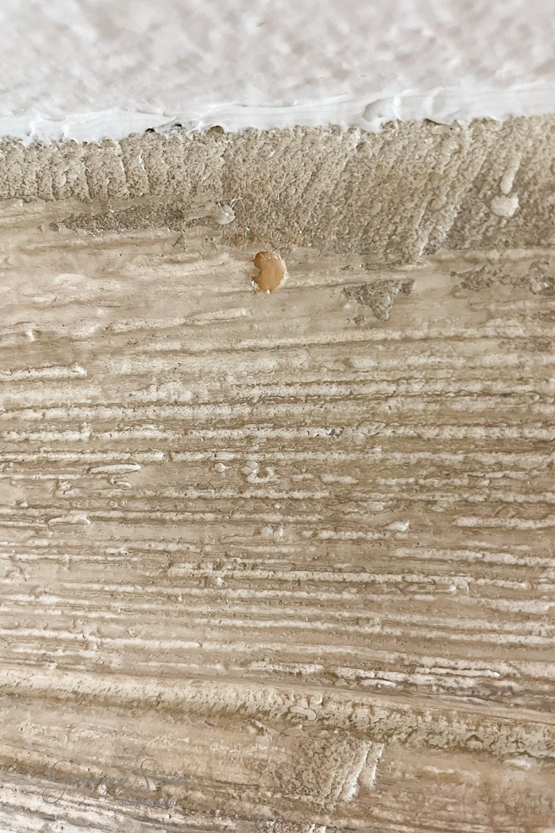 Close-up of a beige textured wall with rough horizontal lines and a small patch of brown paint. The upper edge meets a white ceiling, highlighting the slight unevenness in the wall texture.