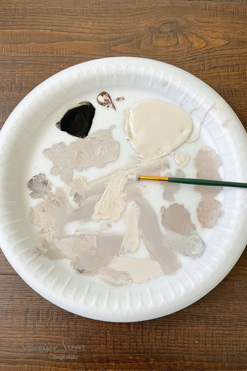 A white disposable plate with various shades of beige, brown, and black paint sits on a wooden surface. A small paintbrush rests partly in one of the beige paint blobs.