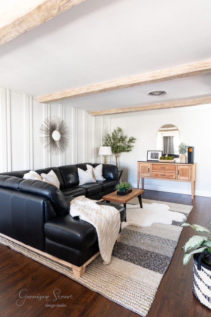 A cozy living room with a black leather sectional sofa adorned with white pillows and a knitted throw. A wooden console table with decor items rests against the wall. The space features DIY faux ceiling beams, sunburst wall art, a soft rug, and elegantly crafted elements throughout.