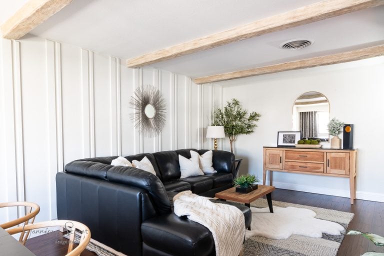 A cozy living room features a black leather sectional adorned with white pillows and a knitted throw. A wooden table and console add warmth, complemented by a circular mirror and sunburst wall decor. DIY faux ceiling beams enhance the white paneled walls, creating an inviting ambiance.