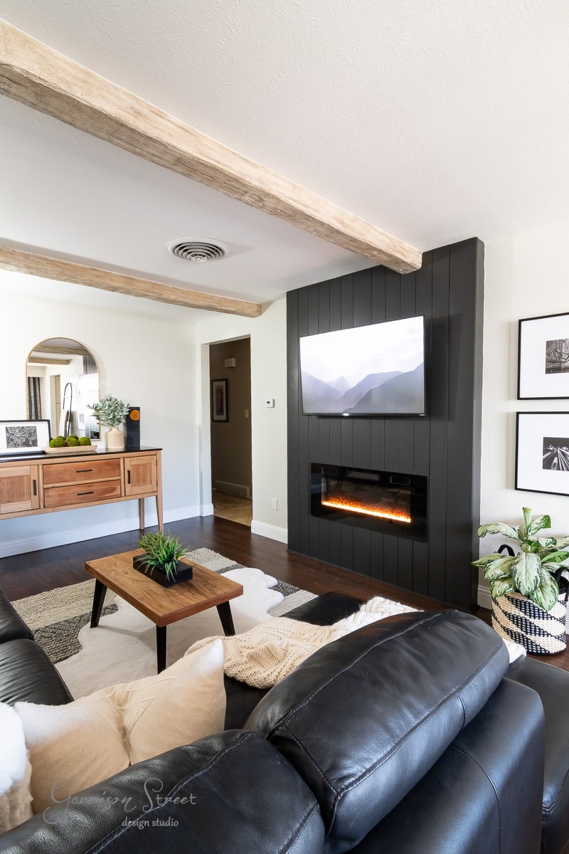 A modern living room featuring a black leather sofa, a wooden coffee table with a small plant, and a wall-mounted TV above an electric fireplace. The room boasts a light wood sideboard and framed photos on the wall.