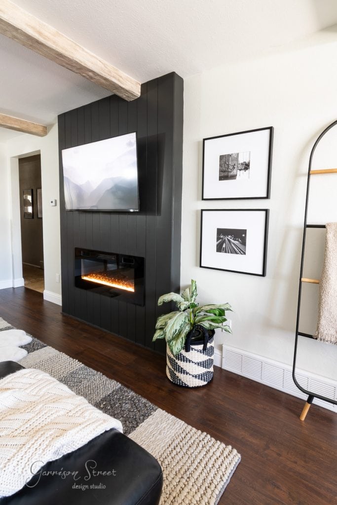 A modern living room with a dark accent wall featuring a mounted TV and electric fireplace. Two black and white photos hang beside it, complemented by a potted plant and blanket ladder. The space is elevated with DIY faux ceiling beams adding rustic charm to the decor.