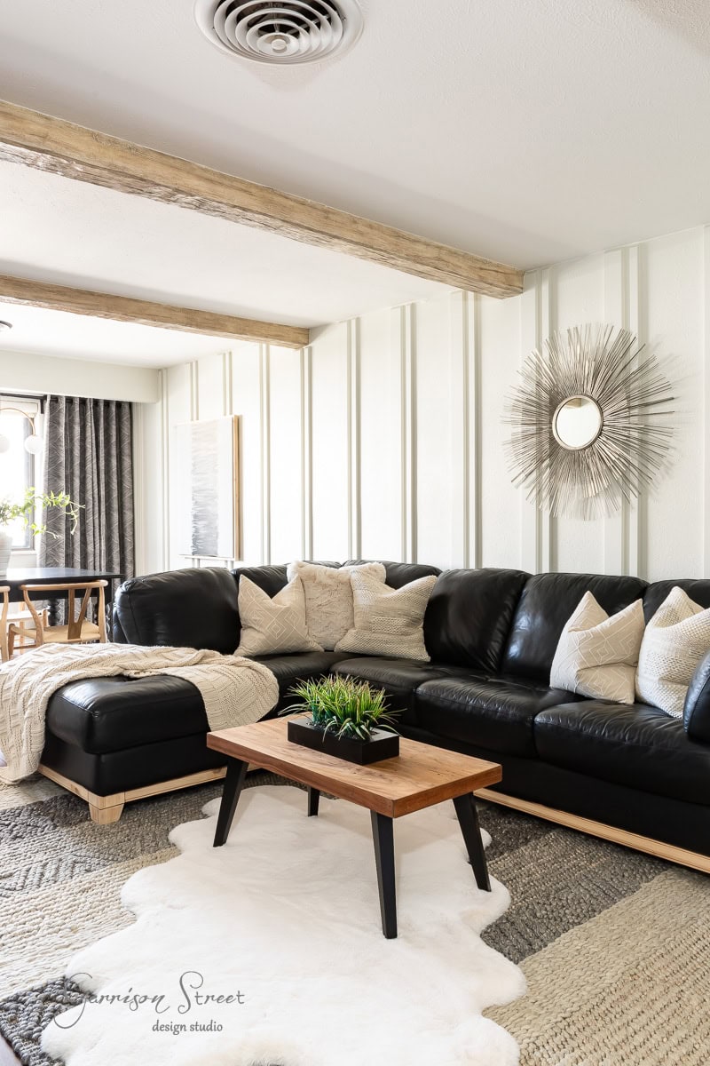 A cozy living room features a black sectional sofa adorned with beige pillows and a knit throw. A wooden coffee table with a plant sits on a white rug. Walls are enhanced with DIY faux ceiling beams and a sunburst mirror, while a dining area is visible in the background.