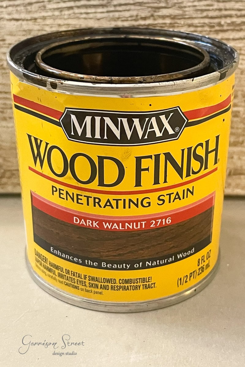 A can of Minwax Wood Finish, Dark Walnut 2716, is shown. The label describes it as a penetrating stain enhancing the beauty of natural wood. The can contains 8 fl oz (1/2 pt) and includes safety warnings.