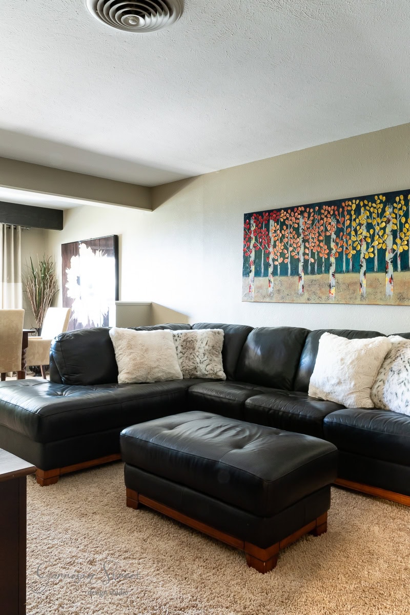 A spacious living room with a black leather sectional sofa and matching ottoman on a plush beige carpet. The wall showcases colorful tree-themed artwork, while a dining area with chairs and a table is visible in the background.