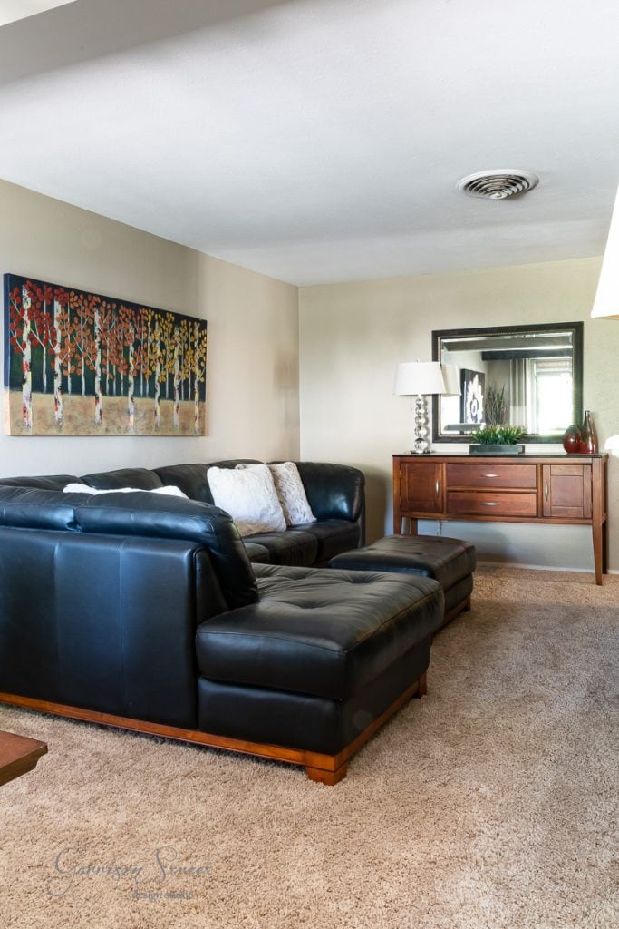 A cozy living room features a black leather sectional sofa adorned with two white pillows and a matching ottoman on beige carpet. A colorful tree painting graces the wall, while DIY faux ceiling beams add rustic charm. A wooden console table with a lamp and mirror completes the scene.