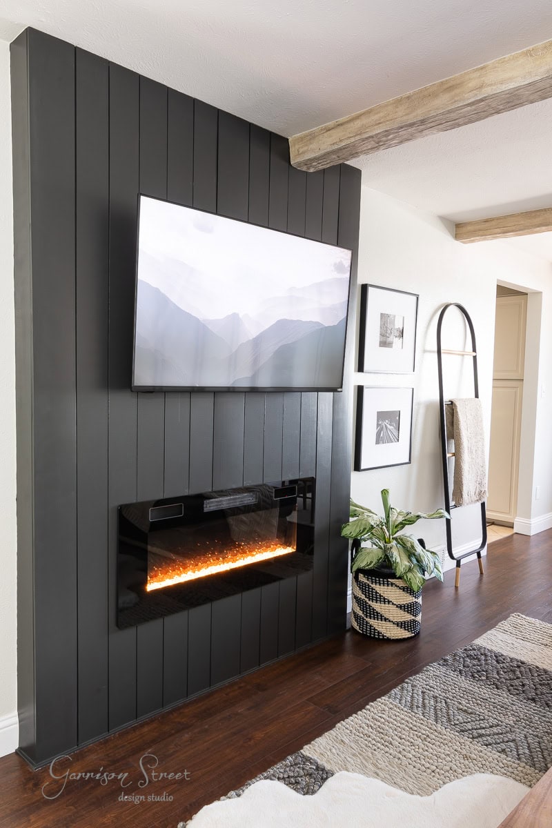 A modern living room boasts a sleek, black shiplap accent wall with a mounted TV above an electric fireplace. Beams add character overhead, complementing the wooden floors, layered rug, ladder shelf with blankets, framed pictures, and a plant in a woven basket.