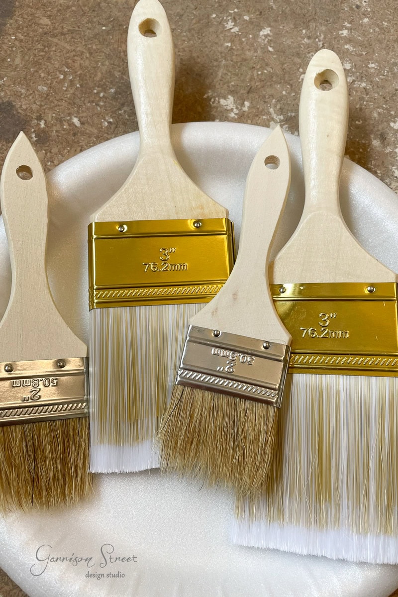 Four new paintbrushes with wooden handles and gold metal ferrules rest on a white circular surface. The bristles are clean and neatly arranged, with sizes labeled on each brush. Against a light brown textured backdrop.