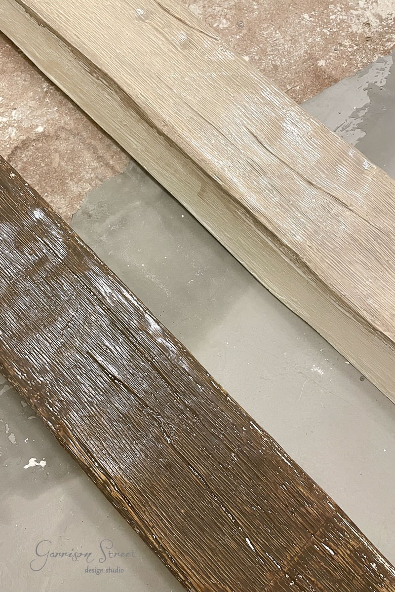 Two wooden beams with a textured finish, one light and one dark, are placed side by side on a concrete surface. The light surface beneath them is partially covered in a thin layer of paint or dust.