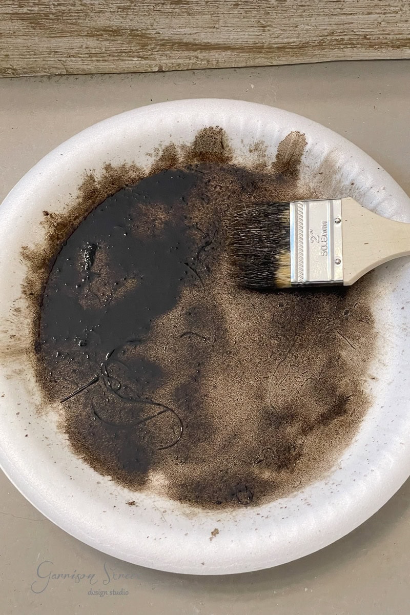A paintbrush rests on a white paper plate covered in dark brown paint and powdery residue. The plate is set against a neutral surface.